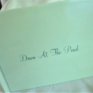 May include: A light green book with the title "Down At The Pond" in elegant script. The book has a glossy finish and is slightly angled, revealing its spine and cover. The background is a soft, white fabric.