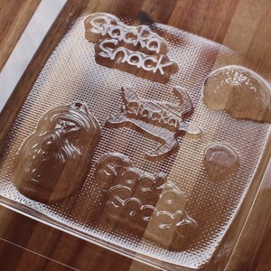 Op de afbeelding: Transparante plastic mal met reliëfvormen voor het maken van chocolade. De mal heeft een schildpad, druiven, een hart en de woorden "Snacka Snack".