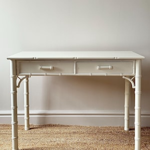 Puede incluir: Un escritorio color crema con dos cajones y patas de estilo bambú. El escritorio tiene una parte superior rectangular y dos cajones con pequeños tiradores rectangulares. Las patas están diseñadas para parecerse a tallos de bambú. El escritorio está sobre una alfombra tejida.