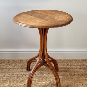 May include: A small, round wooden side table with a light brown finish. The tabletop is approximately 24 inches in diameter. The table has a unique base with curved legs, giving it a vintage or antique appearance. The table is set on a woven rug.