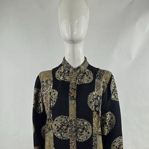 May include: Black and gold patterned dress with a mandarin collar and long sleeves. The dress features an intricate design of gold floral and geometric shapes on a black background. The dress is displayed on a mannequin.
