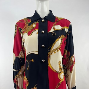 Op de afbeelding: Een lang mouw overhemd met knopen en een geometrisch patroon in zwart, rood en wit met gouden kettingen. Het overhemd heeft een kraag en gouden knopen.