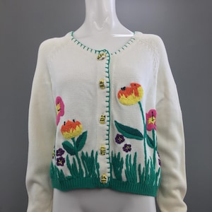 May include: White knitted cardigan with embroidered flowers and bees. The cardigan has a green trim and buttons.