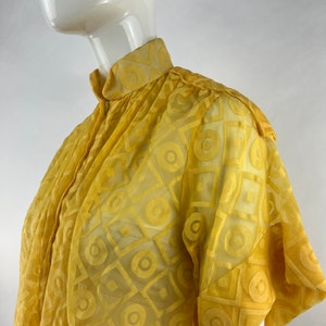 May include: A yellow blouse with a geometric pattern. The blouse has a high collar and short sleeves.