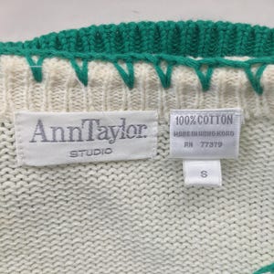 May include: White knitted sweater with a green trim. The sweater has a label that reads "Ann Taylor Studio" and another label that reads "100% Cotton Made in Hong Kong RN 77379 S".