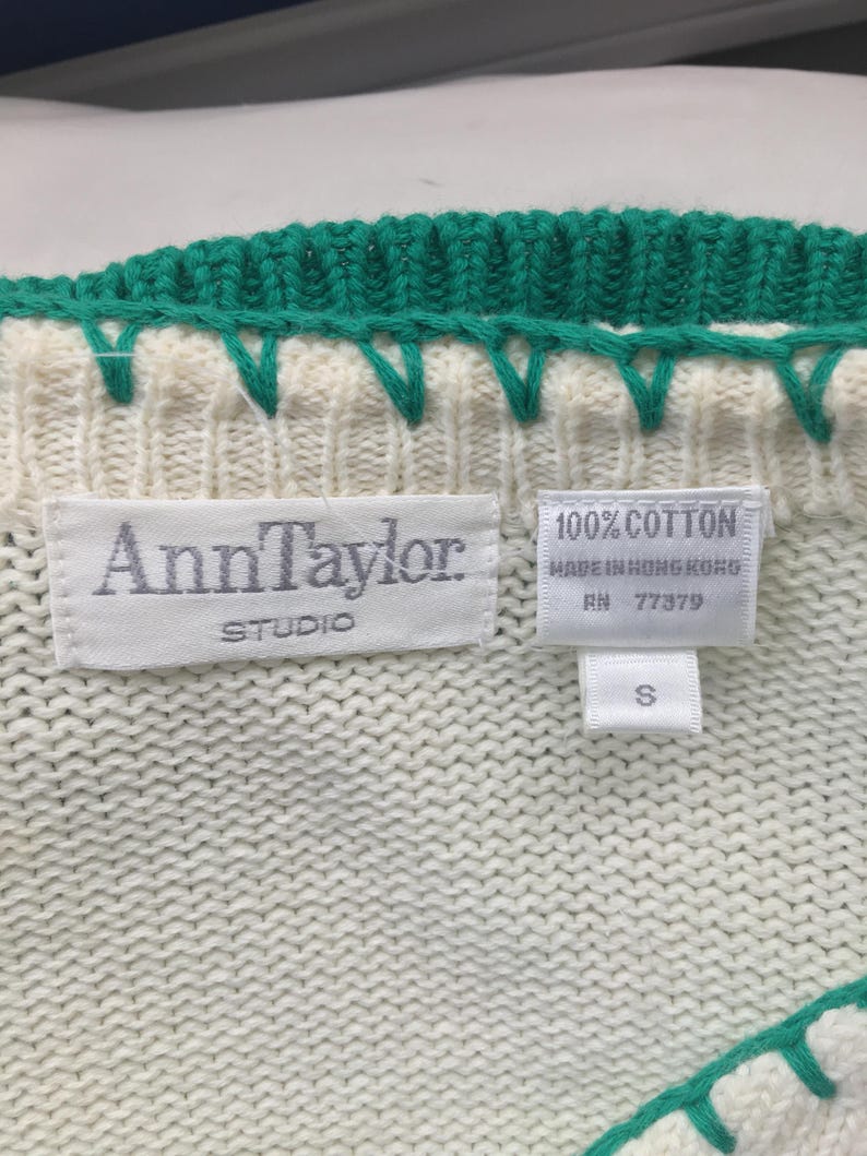 May include: White knitted sweater with a green trim. The sweater has a label that reads "Ann Taylor Studio" and another label that reads "100% Cotton Made in Hong Kong RN 77379 S".