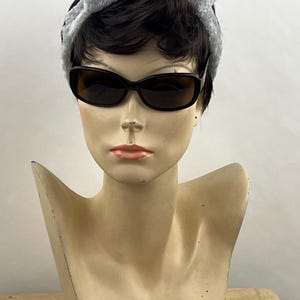 May include: A mannequin head with short dark hair wearing a light gray plush knotted headband and black-framed sunglasses with dark tinted lenses. The mannequin has a neutral facial expression with light skin tone and peach-colored lips.