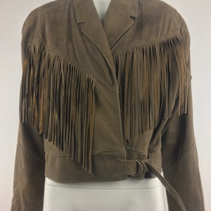 May include: A brown suede jacket with fringe detailing around the collar and hem. The jacket has a belt with a metal buckle.