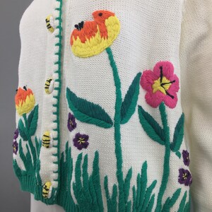 May include: White knitted cardigan with embroidered flowers, bees, and green grass. The cardigan has a button closure.