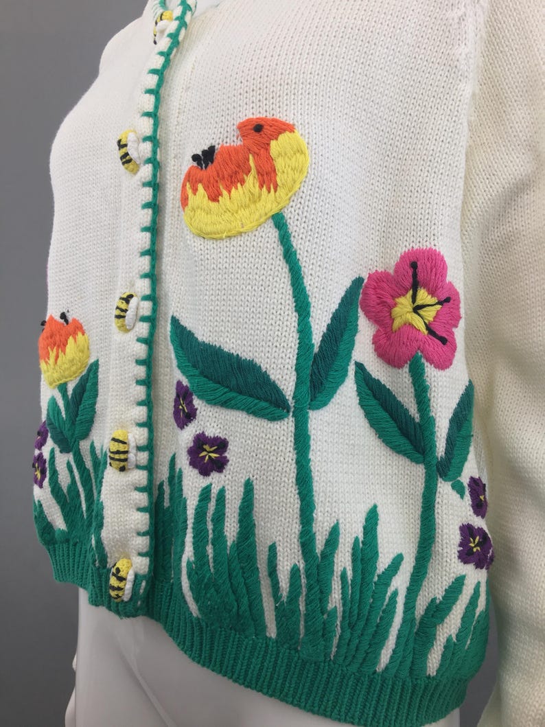 May include: White knitted cardigan with embroidered flowers, bees, and green grass. The cardigan has a button closure.