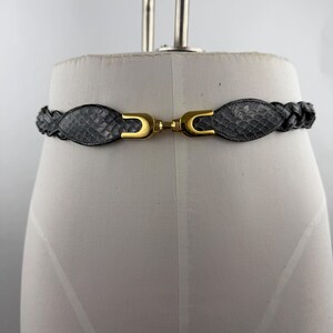 May include: A braided belt featuring a grey snakeskin-patterned texture and a gold-toned metal hook clasp. The belt consists of two oval-shaped snakeskin-textured panels connected by a central gold-toned metal fastener, with braided sections extending outward.
