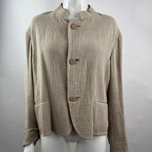 May include: A beige textured jacket with a mandarin collar and three large round buttons down the front. The jacket features long sleeves and two horizontal welt pockets at the waist, crafted from a fabric with a visible crinkled weave pattern.