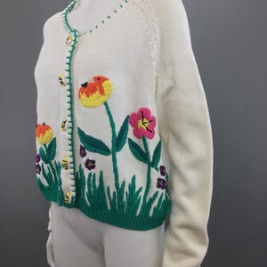 May include: A white knitted cardigan with green, yellow, pink, and purple embroidered flowers and bees. The cardigan has a button closure and a green trim.