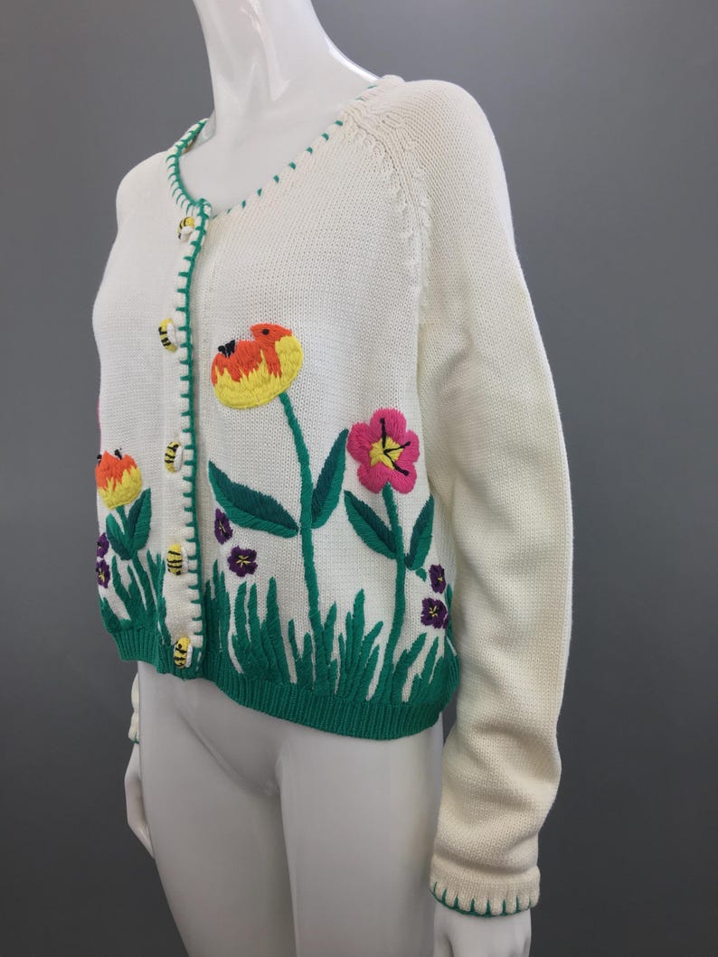 May include: A white knitted cardigan with green, yellow, pink, and purple embroidered flowers and bees. The cardigan has a button closure and a green trim.