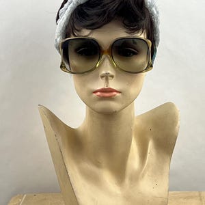May include: A mannequin head wearing a light blue textured headband and oversized sunglasses with a brown gradient tint and a translucent amber frame. The mannequin has short dark brown hair and a neutral facial expression with peach lips.