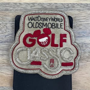 May include: A silver and red embroidered patch with the text "Walt Disney World Oldsmobile Golf Classic".