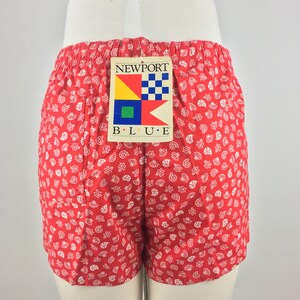 May include: Red shorts with a white seashell pattern. The shorts have a tag that says "NEWPORT B.L.U.E." with a nautical flag design.