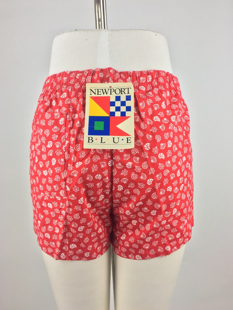 May include: Red shorts with a white seashell pattern. The shorts have a tag that says "NEWPORT B.L.U.E." with a nautical flag design.