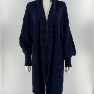 May include: A navy blue open-front cardigan made of a textured, fuzzy knit fabric. This long-sleeved garment features a wide ribbed shawl collar, dropped shoulders, and ribbed cuffs. The design includes two front pockets and a mid-thigh length.