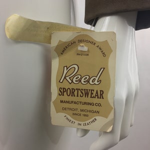 May include: A brown leather tag with the text "American Designer Award" and "Reed Sportswear Manufacturing Co. Detroit, Michigan Since 1950 Finest in Leather".