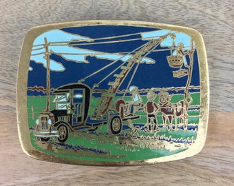 Op de afbeelding: Een gouden gesp met een blauw, groen en wit emaille ontwerp. Het ontwerp toont een vrachtwagen met een kraan, twee figuren en een koe.