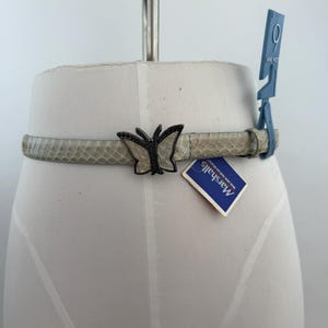 May include: A slim, light grey belt with a textured snakeskin-like pattern. The belt features a prominent dark metal butterfly-shaped buckle with small black embellishments. A blue plastic tag with the number 9 and a Marshalls price tag are attached.