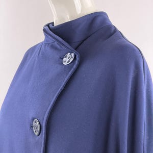 May include: A dark blue coat featuring a high collar and an asymmetrical front closure secured by two large, circular buttons with a distinct geometric pattern. The garment is worn on a mannequin, showcasing the smooth fabric and structured neckline.