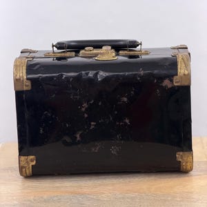 Puede incluir: Un maletín de aseo rectangular negro con acabado brillante, que presenta protectores de esquina de metal en tono latón desgastado, un asa superior negra y un mecanismo de cierre central en tono latón. La superficie muestra signos de desgaste.