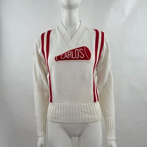 Pode incluir: Camisola creme com decote em V com riscas vermelhas e o nome "CARLOS" em letras vermelhas. A camisola tem mangas compridas e punhos e bainha canelados. Uma camisola desportiva de estilo vintage.