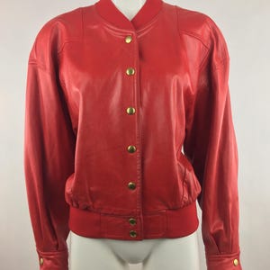 May include: A bright red leather bomber jacket featuring a ribbed collar, long sleeves with buttoned cuffs, and a front closure with six gold-toned metal snap buttons. The jacket has a ribbed hem and structured shoulders, providing a classic silhouette.