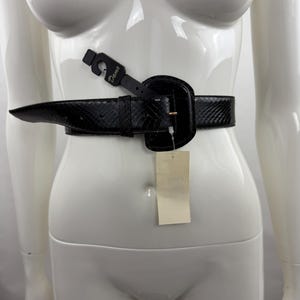May include: A black belt with a snakeskin-like texture and a large, matching black buckle. The belt features a black plastic hanger tag with the word Dame printed on it, and a blank, rectangular beige paper price tag hanging from the buckle.