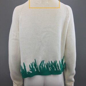 May include: A white knitted sweater with a green trim around the neckline and a green grass design at the bottom.