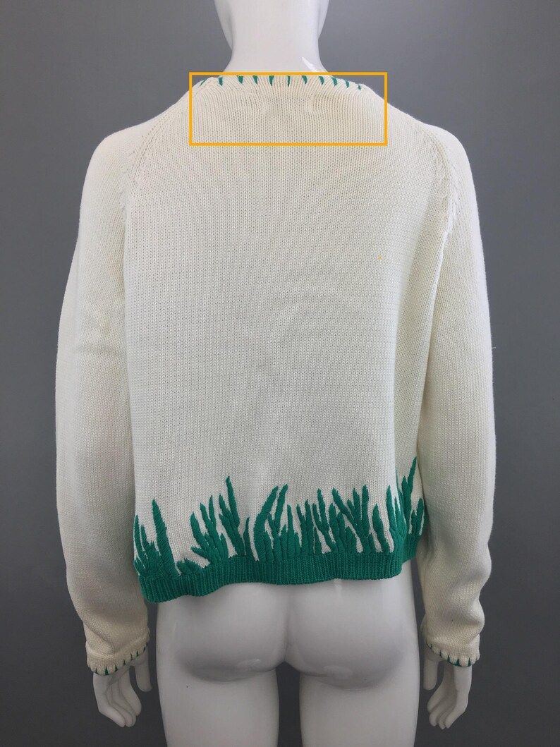 May include: A white knitted sweater with a green trim around the neckline and a green grass design at the bottom.