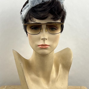 May include: A pair of brown-tinted sunglasses with a thin brown frame and a straight top bar, worn by a mannequin head featuring short dark hair and a light blue textured fabric headband with a knotted bow detail at the top.