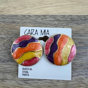 May include: A pair of round, colorful earrings with a swirl pattern in shades of orange, yellow, purple, and pink. The earrings have gold-toned metal settings and surgical steel posts. The earrings are displayed on a white card with the brand name "CARA MIA" printed on it.