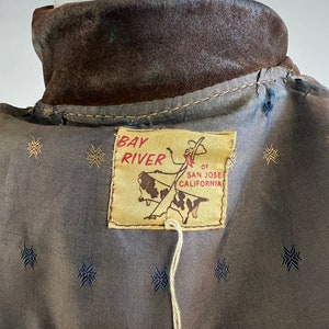May include: Brown suede jacket with a Bay River label. The label features a cowboy riding a bull and the text "Bay River, of San Jose, California".