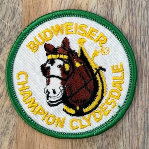 Könnte beinhalten: Ein runder, bestickter Aufnäher mit einem braun-weißen Clydesdale-Pferdekopf in der Mitte. Der Text BUDWEISER CHAMPION CLYDESDALE ist in Gelb um das Pferd herum gestickt. Der Aufnäher hat einen weißen Hintergrund und einen grünen Rand.