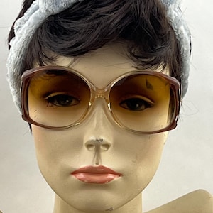 May include: Oversized sunglasses with a gradient brown tint and a thick, dark brown frame. The glasses are worn by a mannequin head featuring short dark hair and a light blue textured fabric headband that sits across the top of the forehead.