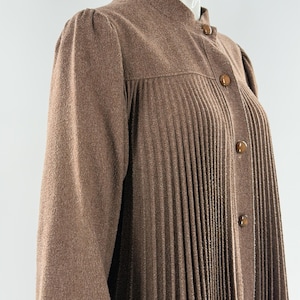 May include: A brown long-sleeved garment featuring a high collar, vertical pleats extending from the chest down, and a front button closure with four round, polished brown buttons. The fabric has a textured, slightly fuzzy appearance.