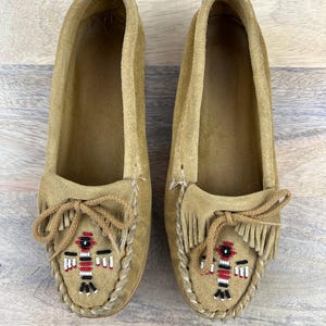May include: A pair of tan suede moccasin-style shoes. Each shoe features a beaded design in red, black, and white, with fringe and a knotted tie detail. The shoes are displayed on a light wood surface.