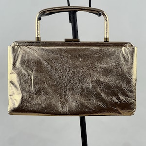 May include: A rectangular metallic gold handbag with a crinkled texture. The bag features a rigid frame, a top-mounted metal clasp closure, and a single arched gold-tone handle attached to the top frame. The surface has a reflective finish.
