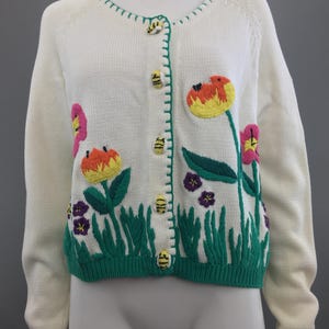 May include: White cardigan sweater with embroidered flowers and bees. The flowers are pink, orange, yellow, and purple. The bees are yellow and black. The cardigan has a green trim around the neckline and cuffs.