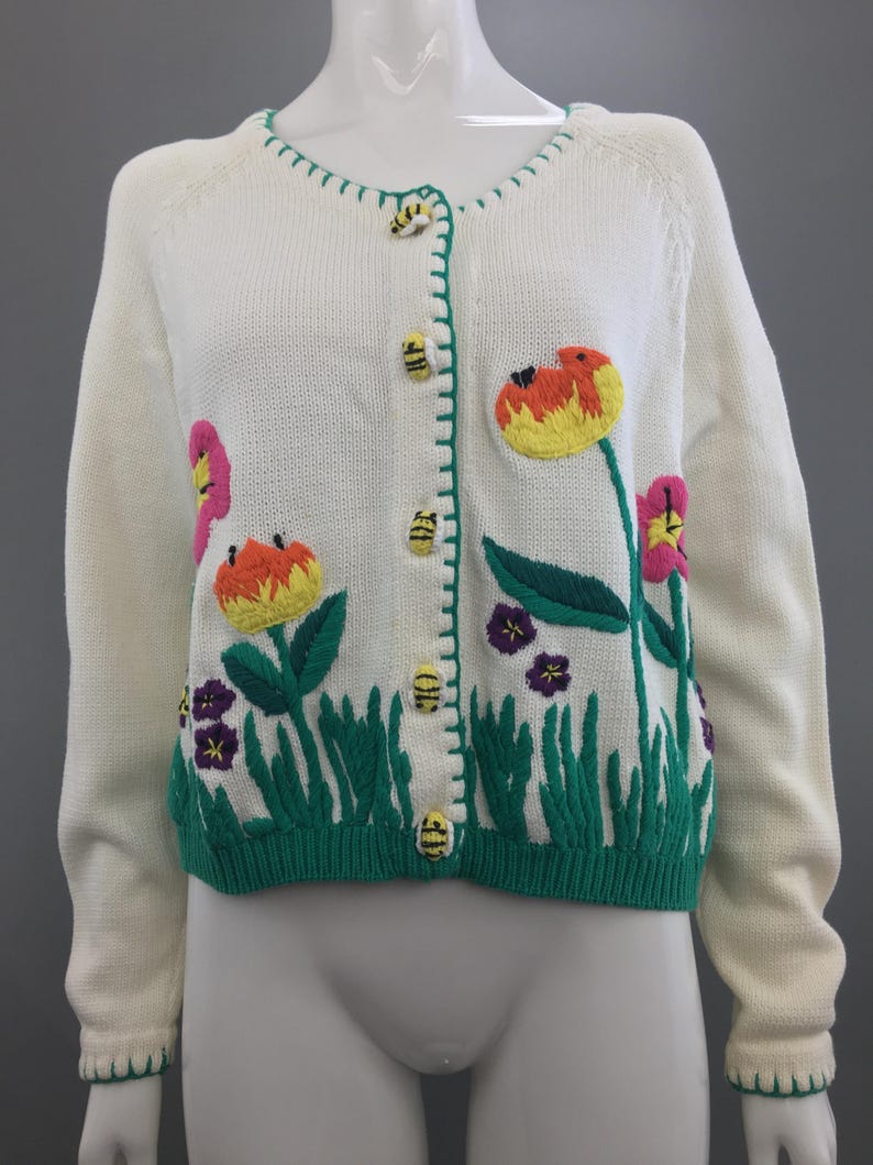 May include: White cardigan sweater with embroidered flowers and bees. The flowers are pink, orange, yellow, and purple. The bees are yellow and black. The cardigan has a green trim around the neckline and cuffs.