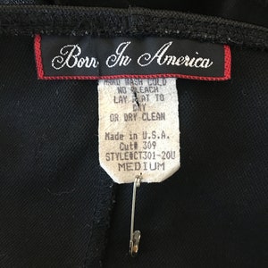 Puede incluir: Una etiqueta blanca con texto negro unida a una prenda negra. La etiqueta dice "Born In America", "Lavar a mano con agua fr&iacute;a", "Sin blanqueador", "Secar en plano", "O limpieza en seco", "Hecho en EE. UU.", "Cut# 309", "Style CT301-200" y "Medium".