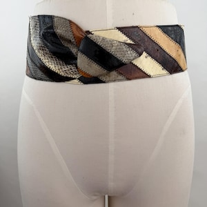May include: A wide waist belt featuring a patchwork design of diagonal stripes in shades of black, brown, cream, and grey. The belt is constructed from textured panels with a snakeskin-like pattern and includes a central knotted detail at the front.