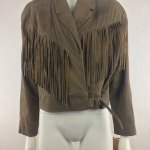 May include: A brown suede jacket with fringe detailing around the collar and hem. The jacket has a belt with a silver buckle.
