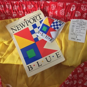 May include: A red and yellow fabric with a white pattern of seashells. The fabric has a tag attached that says "NEWPORT BLUE" and features a colorful geometric design. The tag also includes care instructions and a size label that says "L" and "STYLE 9862".
