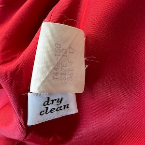 May include: Close-up of a vibrant red fabric with a white tag. The tag reads "dry clean" in cursive and includes size information. The fabric appears to be part of a garment.