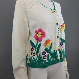 May include: A white knitted cardigan with a green trim and embroidered flowers and bees. The cardigan has a V-neck and button closure.
