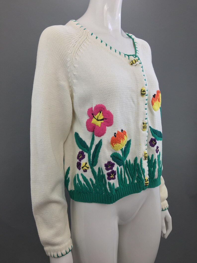 May include: A white knitted cardigan with a green trim and embroidered flowers and bees. The cardigan has a V-neck and button closure.